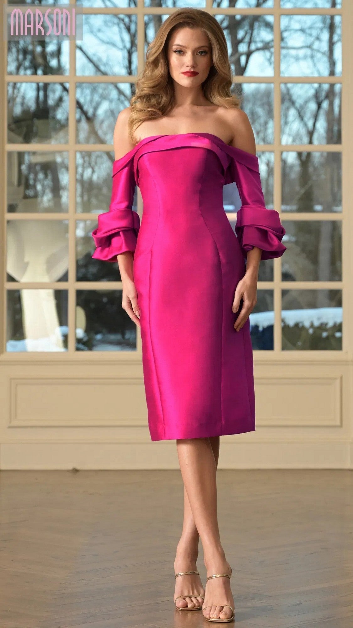 A woman stands indoors before large windows, wearing the Marsoni MV1370 knee-length semi-formal dress in fuchsia with ruffled off-the-shoulder sleeves.